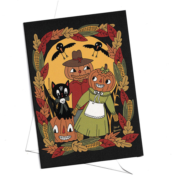 A Halloween-themed art print or card featuring a whimsical illustration of a jack-o'-lantern, a black cat, a scarecrow in a green dress, and crows framed by autumn leaves and corn on a black background, shown against a white backdrop.