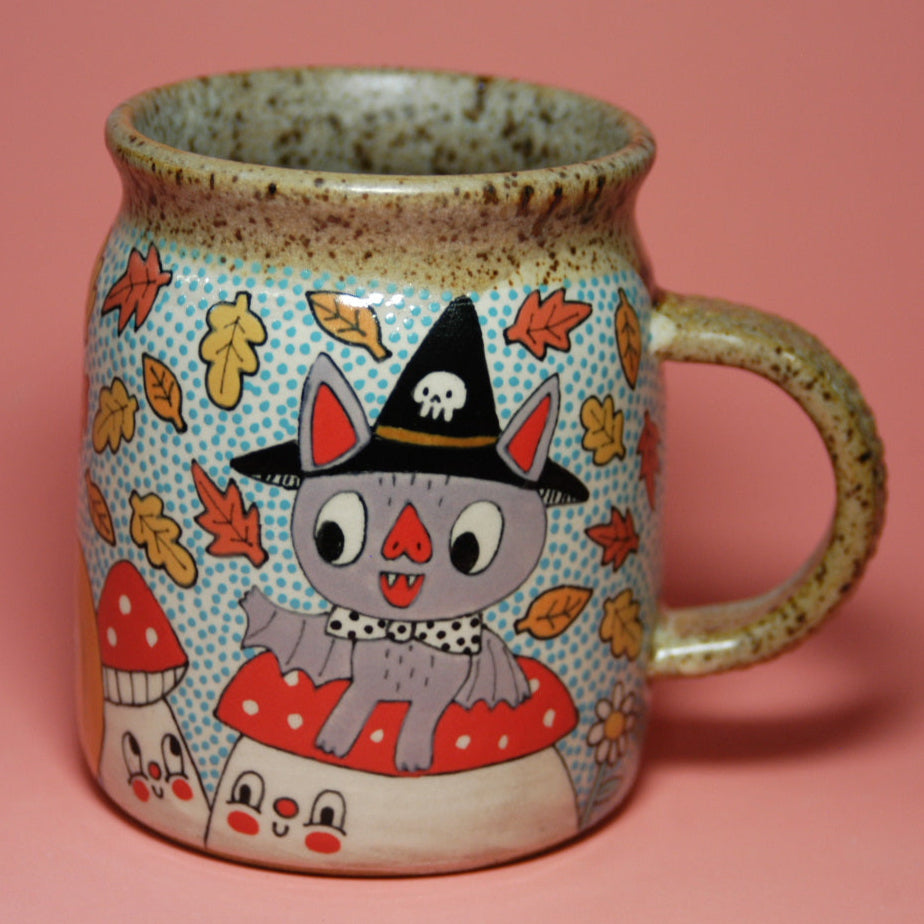 Ceramic mug with a Halloween-themed design featuring a cat in a witch hat and leaves on a pink background.