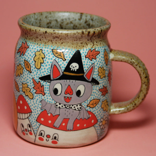 Ceramic mug with a Halloween-themed design featuring a cat in a witch hat and leaves on a pink background.