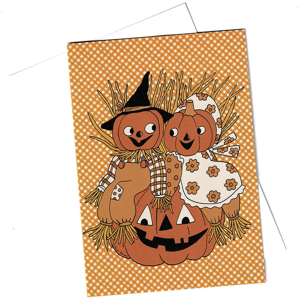 A rectangle Halloween greeting card featuring two scarecrows made from pumpkins, one wearing a black witch hat, and the other in a white dress with orange flowers. They are sitting on a carved pumpkin against an orange-and-white gingham background.