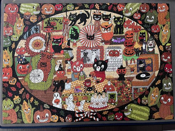 A  jigsaw puzzle featuring a dense, whimsical Halloween illustration with cats, pumpkins, owls, ghosts, and autumn motifs, photographed flat against a dark background.