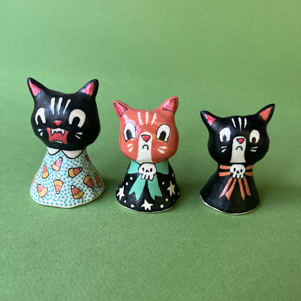 Three hand-painted ceramic cat figurines in black, orange, and black with colorful patterned dresses, arranged on a solid green background.