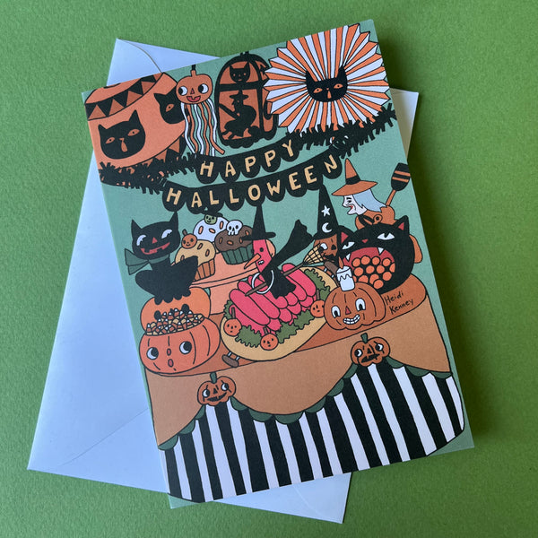 A Halloween greeting card with a whimsical illustration of pumpkins, black cats, witches, and treats on a mint green background, shown with a white envelope.