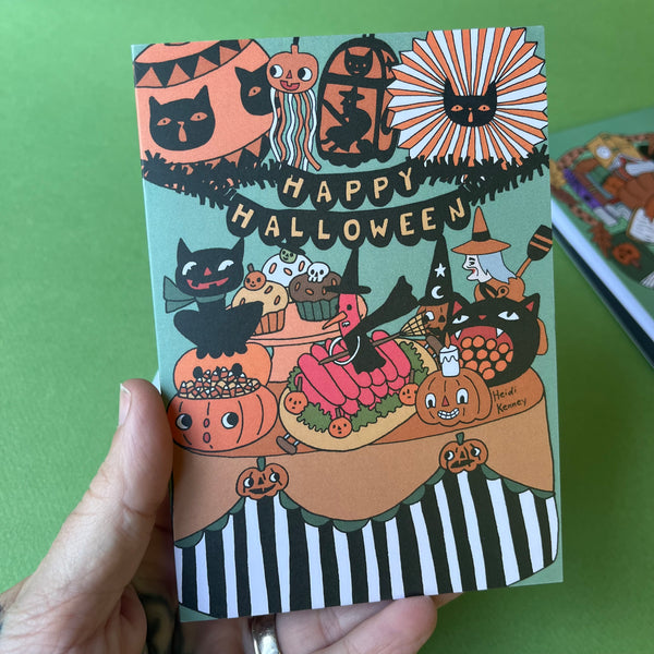A hand holding a Halloween-themed art print or card featuring whimsical illustrations of pumpkins, cats, witches, and the text 'Happy Halloween' on a mint green background, with a green surface behind it.