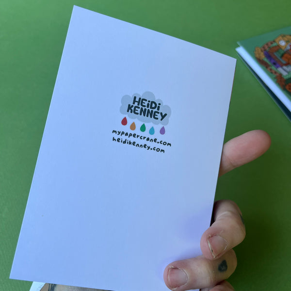 A hand holding a white greeting card with the printed name 'HEIDI KENNEY' and website text, against a green background.