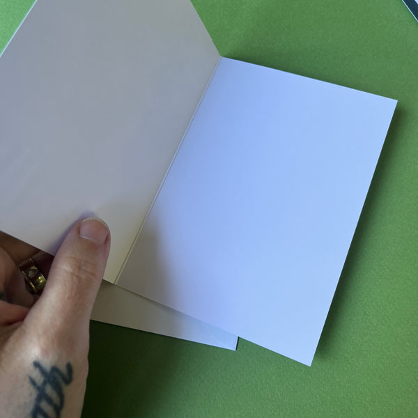 A hand holds an open blank greeting card  against a solid green background.