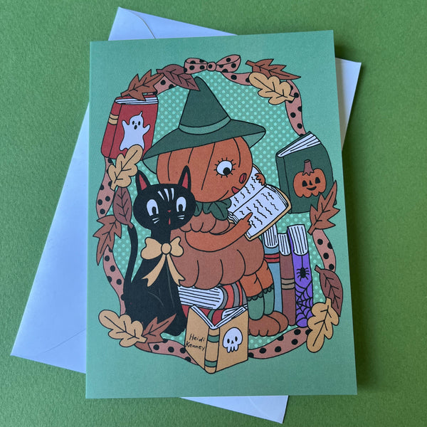 A Halloween greeting card featuring an illustration of a pumpkin character in a witch hat reading a book, with a black cat, books, and autumn leaves on a mint green background. The card is displayed on top of light blue envelopes against a solid green surface.