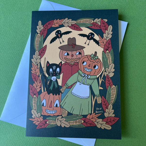 A Halloween-themed art print or greeting card featuring carved pumpkins, a cat , a scarecrow, crows, corn, and autumn leaves, photographed on top of a light blue envelope against a green background.