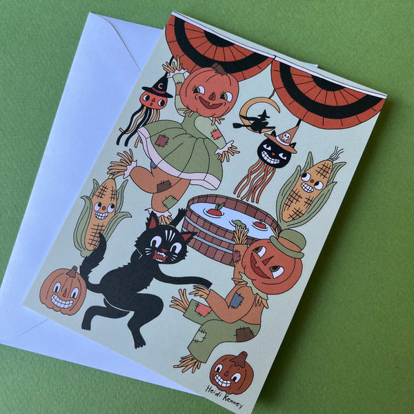 A Halloween-themed greeting card with a whimsical illustration of pumpkins, corn, a black cat, scarecrows, and a witch on an off-white background, shown with a white envelope on a green surface.