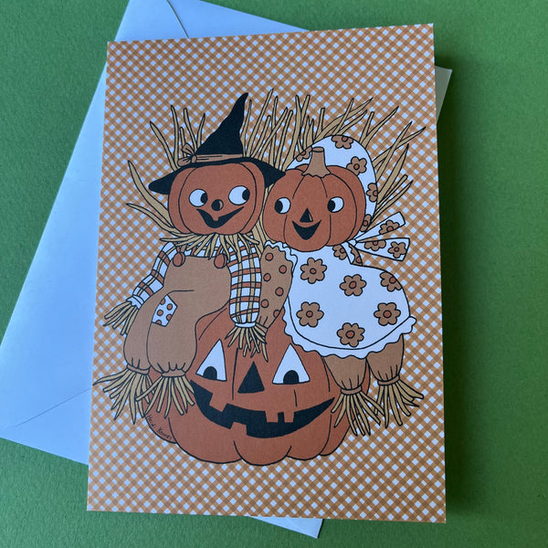 A rectangle Halloween greeting card featuring two scarecrows made from pumpkins, one wearing a black witch hat, and the other in a white dress with orange flowers. They are sitting on a carved pumpkin against an orange-and-white gingham background. on a green background