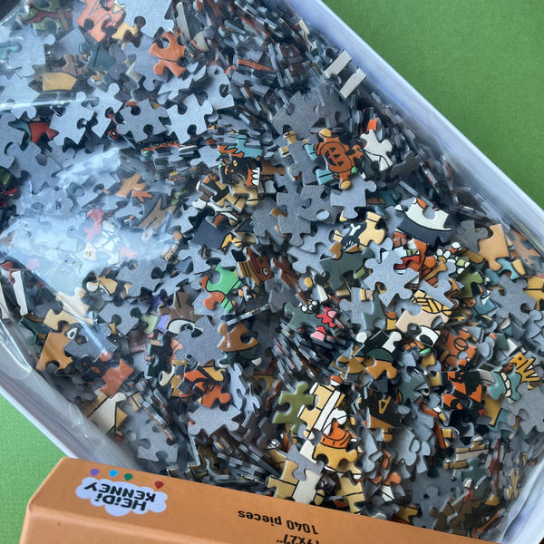 A close-up of a jigsaw puzzle box showing many loose puzzle pieces inside a clear plastic bag. The pieces are multicolored with cartoon-style illustrations.