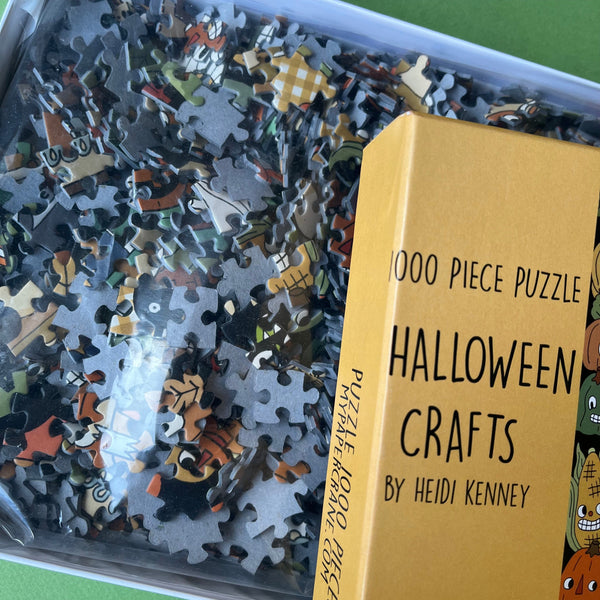 A close-up of a 1000-piece Halloween-themed jigsaw puzzle box by Heidi Kenney, showing the yellow packaging and a clear bag filled with scattered puzzle pieces.