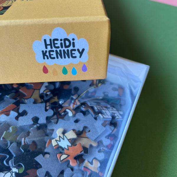 A close-up of a jigsaw puzzle box lid labeled 'HEIDI KENNEY' in a blue cloud, with many puzzle pieces visible inside a clear bag on a green surface.