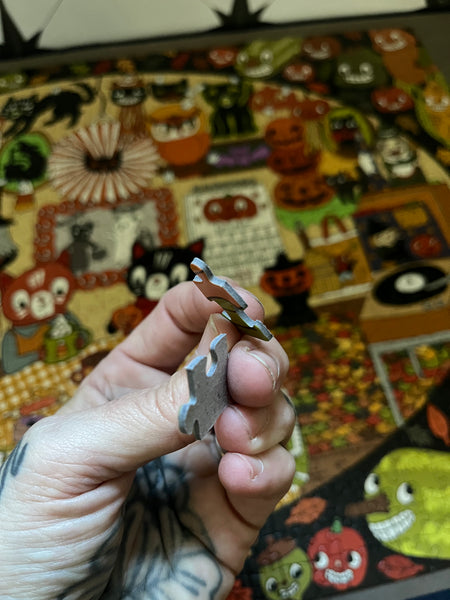 A hand holds a small puzzle piece in front of a colorful, busy Halloween-themed puzzle or craft board with pumpkins, ghosts, and autumn illustrations.