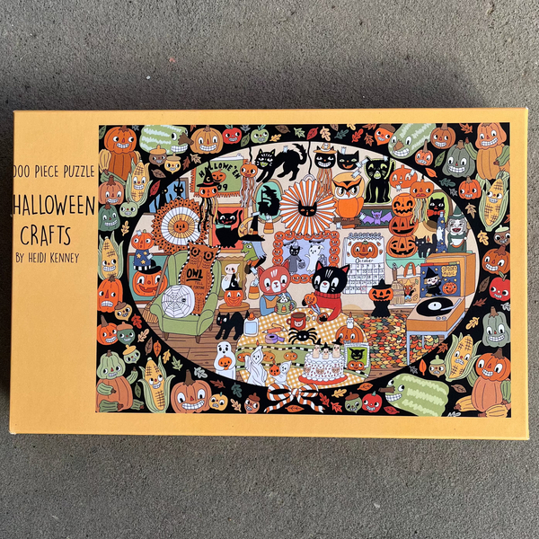 A 1000-piece puzzle box titled 'Halloween Crafts' by artist Heidi Kenney, featuring a colorful illustrated cover with various spooky and festive motifs, photographed flat on a gray concrete surface.