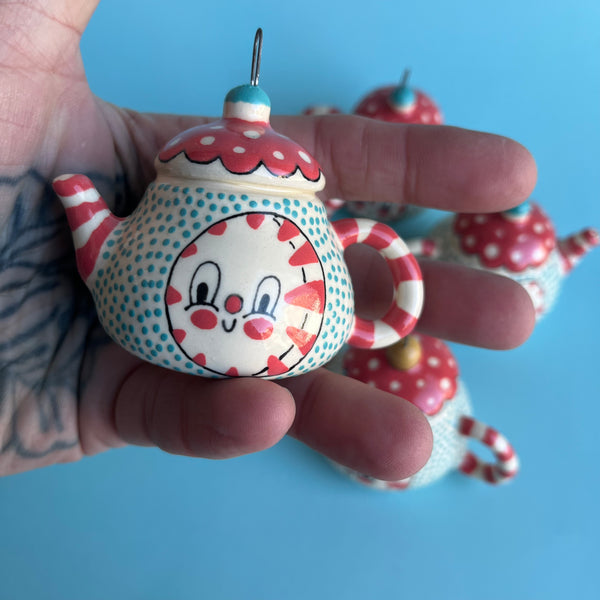 Colorful ceramic teapot-shaped ornament with a peppermint with face held in a hand against a blue background