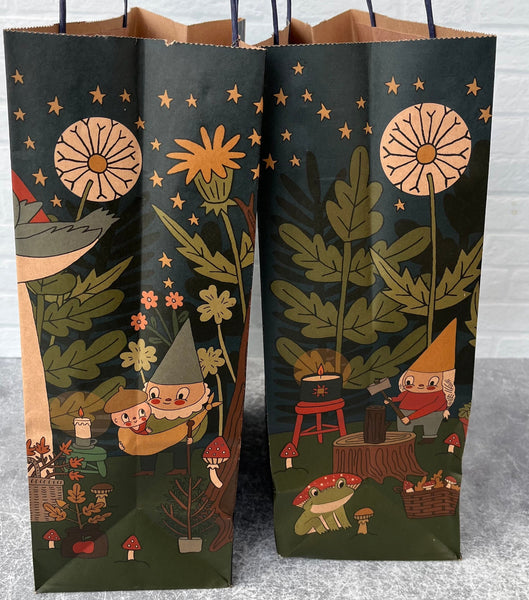 Two paper bags with forest-themed designs featuring gnomes and plants on a gray surface.