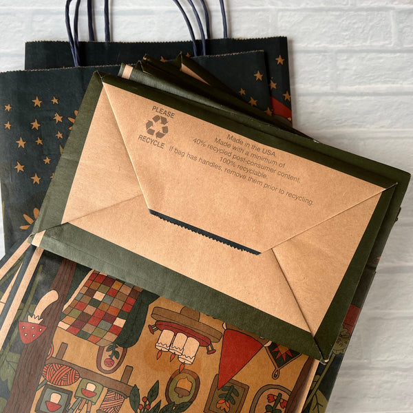 Stack of gift bags with bottom text that reads "made in the USA Made with a minimum of 40% post-consumer content 100% recyclable If bag has handles remove them prior to recycling Please recycle" on a white brick background