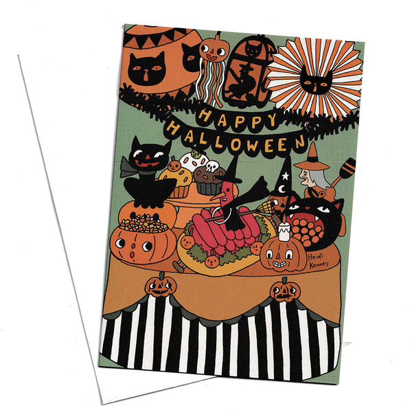 A Halloween-themed greeting card with a whimsical illustration of pumpkins, black cats, witches, cupcakes, and the words “HAPPY HALLOWEEN” on a pale green background, shown with a white envelope on a white surface.