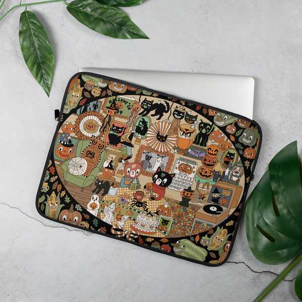 A laptop sleeve with a detailed, colorful Halloween-themed collage print featuring cats, pumpkins, ghosts, and autumn motifs, shown on a silver laptop against a light gray textured background with green leaves.