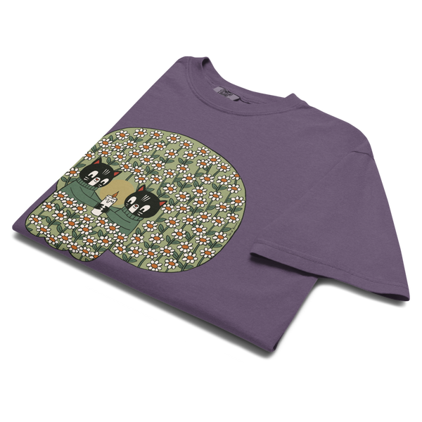 A folded purple crewneck t-shirt with a large graphic print on the front. The graphic features a green skull patterned with small white and flowers, containing two cartoon cats.