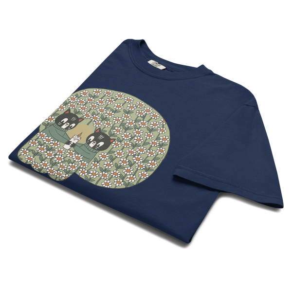 A navy blue short-sleeve t-shirt with a large graphic print on the front. The graphic features two cartoon cats in green sweaters and white flowers on an green skull shaped background.